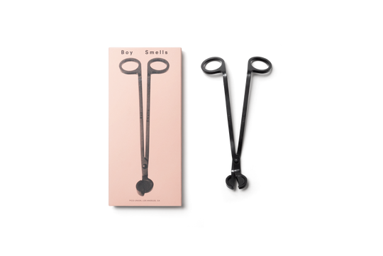 product shot of exterior pink box with black text, boy smells logo in black and black image of wick trimmer , next to box is metallic black wick trimmer with engraved boy smells logo against a neutral background.