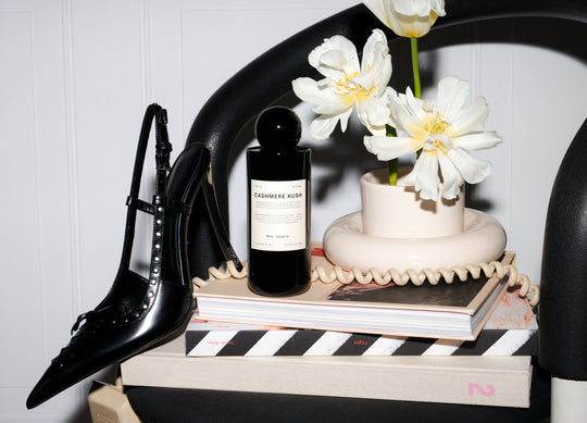Editorial image of Cashmere Kush Room spray sitting on a stack of books on a chair surrounded by a modern white vase and black high heel and a phone cord hanging off edge.