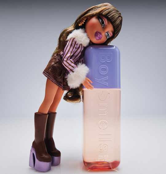 Doll leaning against a bottle of BOY SMELLS perfume with a gray background
