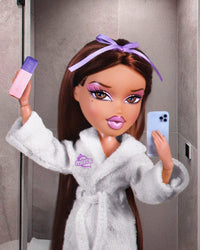 Bratz doll in a white robe with a purple headband holding a phone and a bottle.