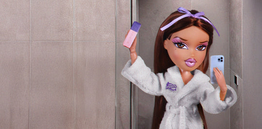 Doll in a white robe with a logo, holding makeup and a phone, in a bathroom setting.